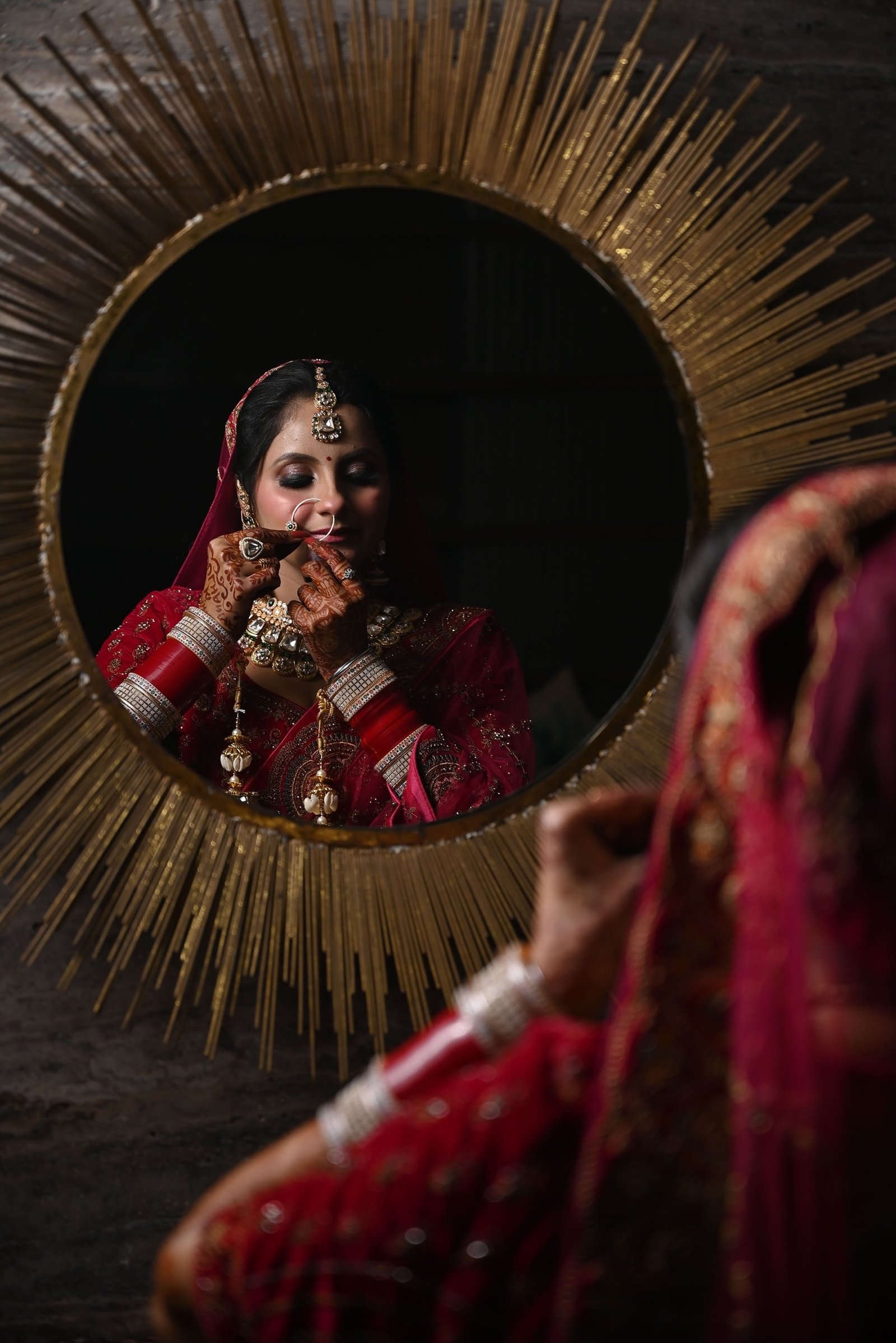 Bride adjusting her nath, reflected in a gold sunburst mirror